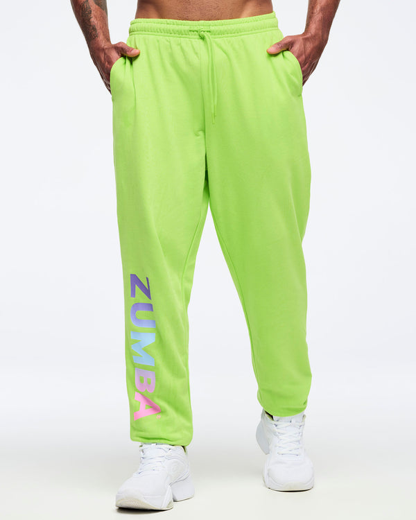 Front view of neon green Zumba joggers with elastic drawstring waistband and relaxed fit, featuring large vertical “ZUMBA” gradient lettering on the lower leg, worn by a male model with white sneakers.
