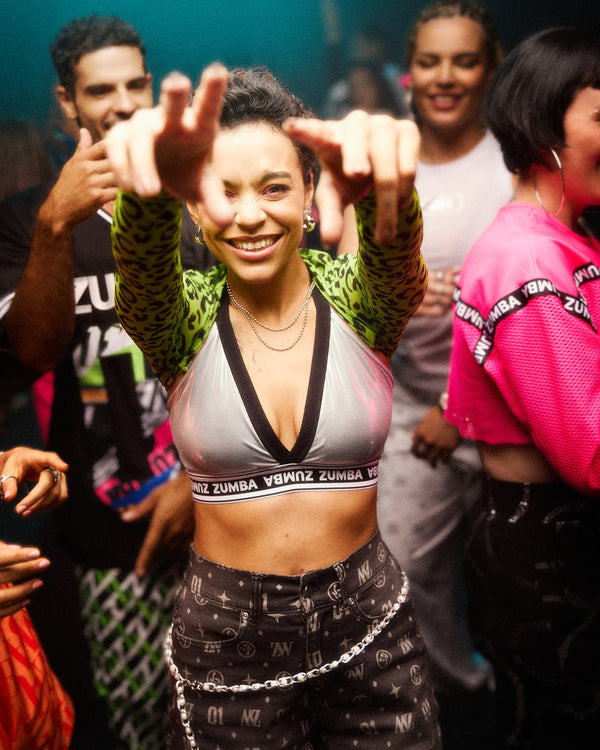 A female model in a dance club wearing a silver and black trimmed Zumba sports V-bra and patterned jorts, posing with her hands in front of her face while surrounded by others.