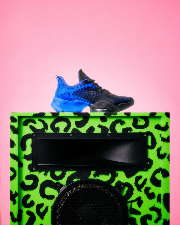 A single athletic sneaker in black and bright blue is placed on top of a green speaker cabinet with a black leopard print, against a solid pink background.