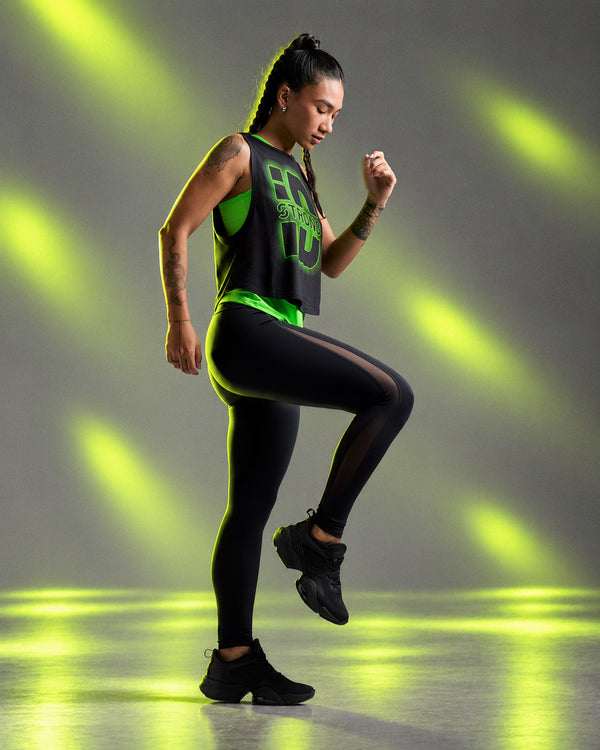 Female model in the black mesh insert leggings, mid-step with one knee raised, wearing a black 'STRONG iD' crop muscle tank over a neon green top. Neon light beams on a grey backdrop.