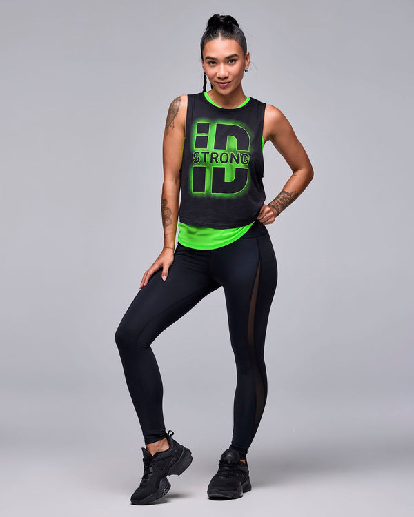 Full view of a female model in the black 'STRONG iD' crop muscle tank layered over a neon green top, with black mesh insert leggings and black shoes.