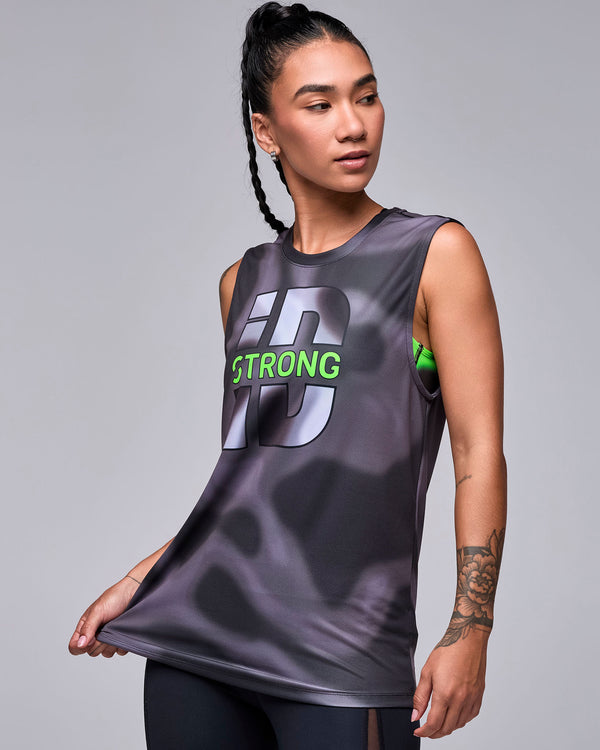 Female model pulling at the hem of the black and grey 'STRONG iD' tank, with a neon green bra visible at the armhole and black mesh insert leggings.