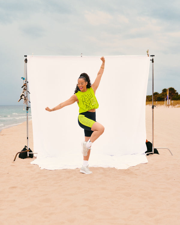 Neon yellow Zumba crop tank and black fitted biker shorts with neon yellow side panels. Female model wears the bright cropped tank with high-waisted shorts, white crew socks, and silver athletic sneakers, posing on sand in front of a white backdrop.