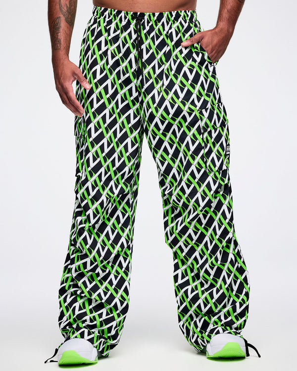 Front view of a male model wearing black cargo pants with an allover black, white and green “Z” crisscross pattern, left hand in his pocket, paired with black-and-white shoes.