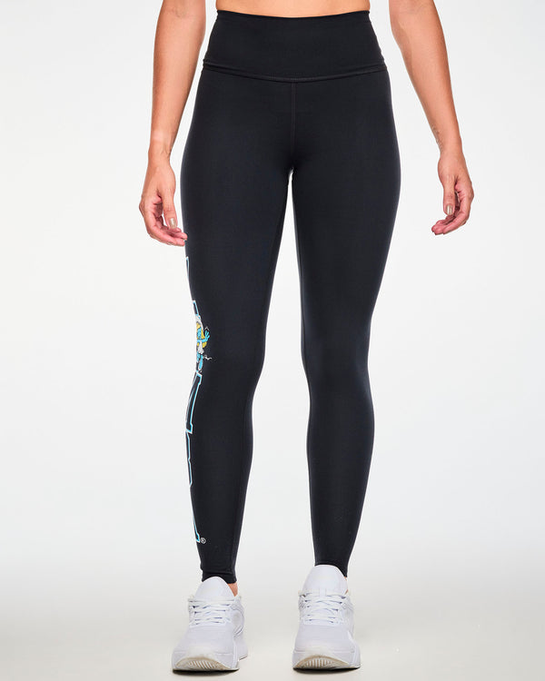 Female model in black high-waisted ankle leggings with vertical varsity 'ZUMBA' text outlined in blue and white running down the outer right leg, barely visible from the front, with Smurfette waving near the top. Paired with white shoes.