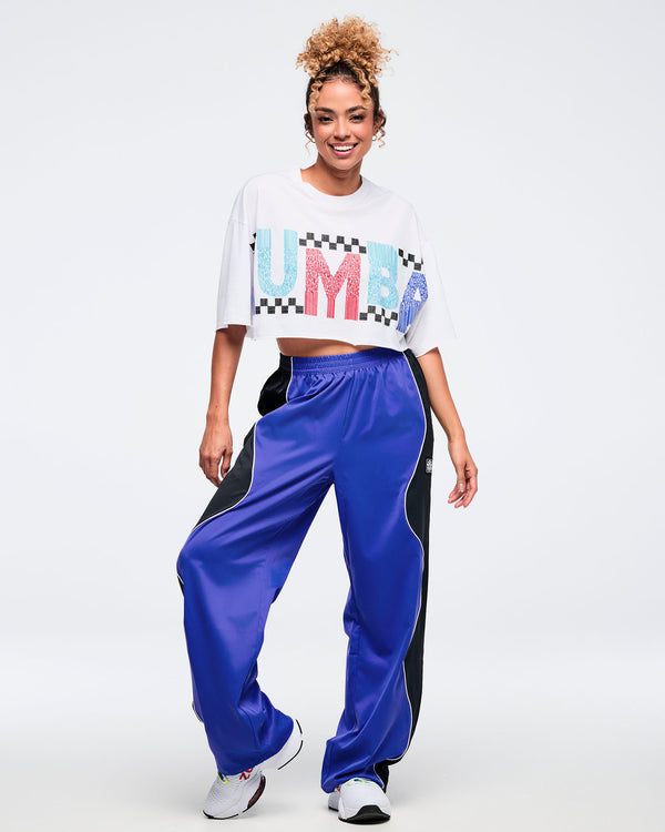 Full-body view of the white cropped oversized graphic tee and shiny royal blue track pants with black side panels and white piping, worn by a female model with white sneakers in a studio setting.