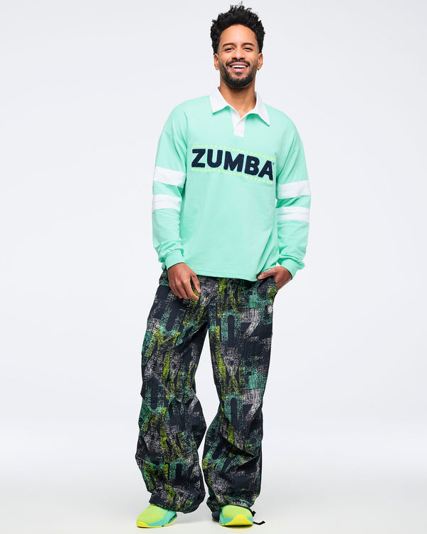 Mint green long-sleeve rugby sweatshirt with white collar, white sleeve stripes, and large navy “ZUMBA” chenille-style chest lettering; male model pairs it with black-and-green abstract print pants and ombre green sneakers on a white background.