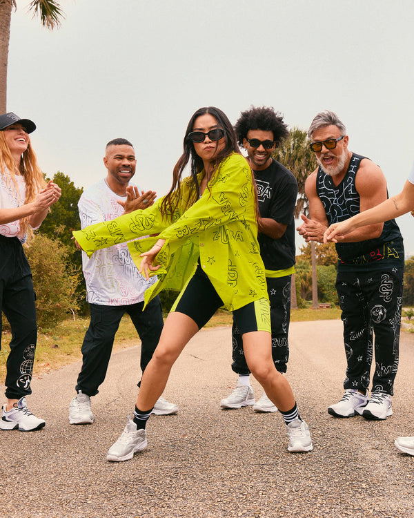 Female model wears the oversized shirt, fitted black shorts, black striped crew socks, silver sneakers, and black sunglasses while dancing on a road with other models in Zumba activewear.