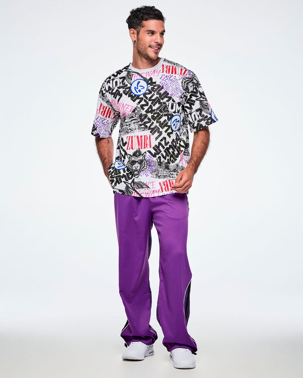 Full view of a male model wearing a graphic tee with black, white, pink, and blue graffiti-style print and logos, such as blue zumbitos, a tiger wearing sunglasses,