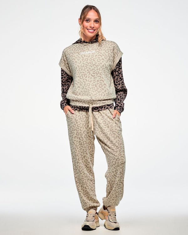 Full view of a female model in a beige leopard print sleeveless sweatshirt layered over a brown leopard print hoodie, paired with matching beige leopard print pants and beige shoes.