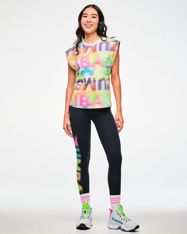 Female model wearing a multicolor halftone print extended shoulder tank, paired with black ZUMBA leggings, pink striped socks, and white sneakers.