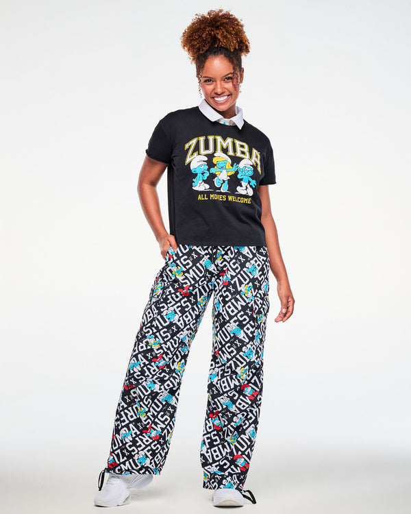 Female model with one hand in her pocket, wearing the black Smurfs graphic top with 'ZUMBA,' three dancing Smurfs, and 'ALL MOVES WELCOME' over a white-collared mesh polo, paired with black and white Smurfs print cargo pants.
