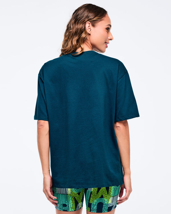 Back view of the dark teal oversized T-shirt on a female model, showing the clean solid back and loose sleeves, paired with green-and-blue patterned biker shorts.
