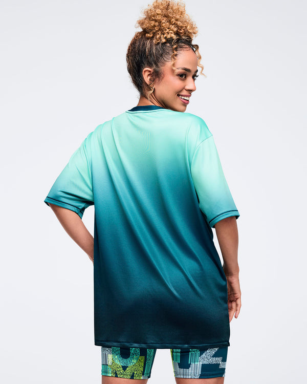Oversized ombre aqua-to-deep teal short-sleeve T-shirt with dark crewneck, shown from the back on a female model; loose fit paired with green-and-teal patterned biker shorts.