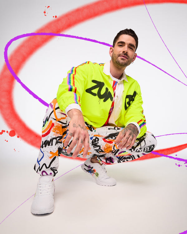 Male model wearing a bright yellow jacket with 'Zumba' black text, colorful pants, and white sneakers on a white background with red and purple abstract designs.