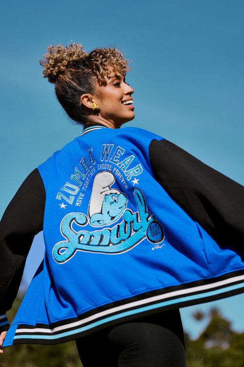 Female model twirling in a blue and black varsity jacket, showing the chenille back graphic of 'ZUMBA WEAR,' a winking Smurf, and script 'Smurf' text, against a clear blue sky.