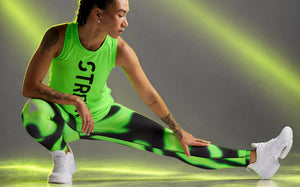 Female model stretching in a side lunge, one hand on her extended leg, wearing a neon green basketball tank with black 'STRONG' text, neon green and black swirl print leggings, and white shoes. Grey background with diagonal neon light beams.