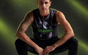 Male model crouching with his hands clasped, in a black and grey swirl print wide shoulder tank with a large grey 'iD' graphic and neon green 'STRONG' across the middle, paired with black pants. Dark background with diagonal neon light beams.