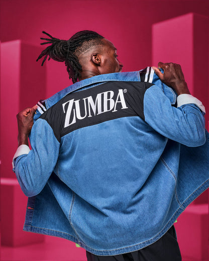 Back view of a male model holding open a denim shirt with white 'ZUMBA' text embroidered on a black yoke, against a pink backdrop.