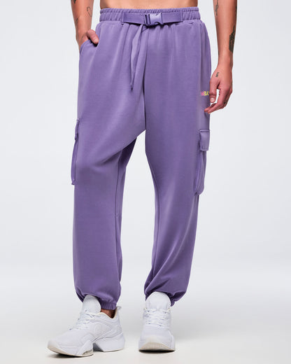 Front view of a male model wearing purple slouch cargo jogger pants with an elastic waistband, adjustable belt, side cargo pockets, and tapered elastic cuffs, paired with white sneakers on a white background.