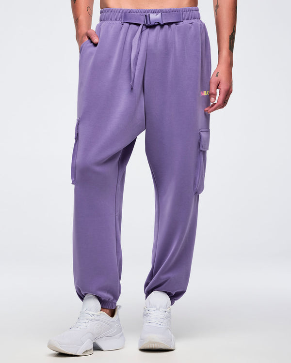 Front view of a male model wearing purple slouch cargo jogger pants with an elastic waistband, adjustable belt, side cargo pockets, and tapered elastic cuffs, paired with white sneakers on a white background.