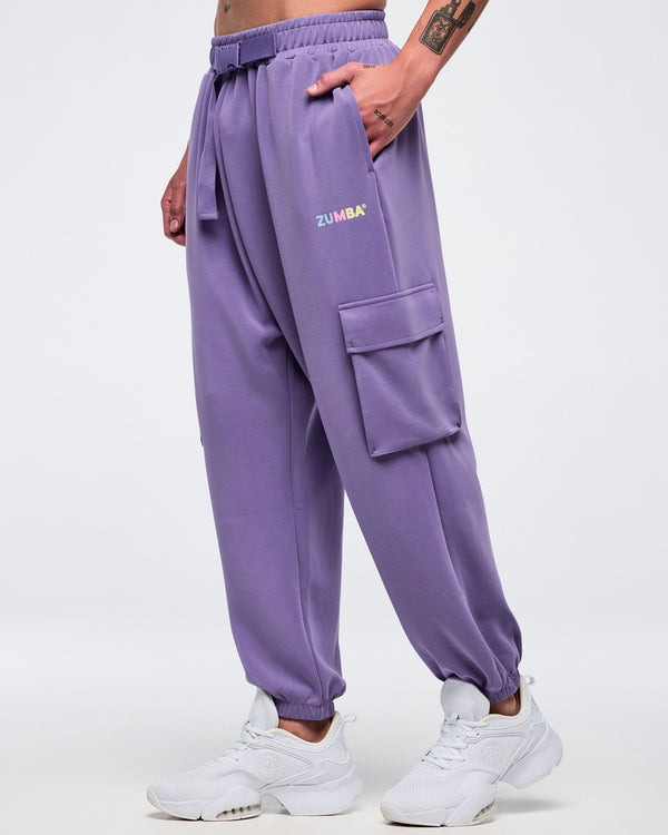 Side view of a male model wearing purple slouch cargo jogger pants with an elastic waistband, adjustable belt, side flap cargo pocket, and a small “Zumba” logo, paired with white sneakers on a white background.