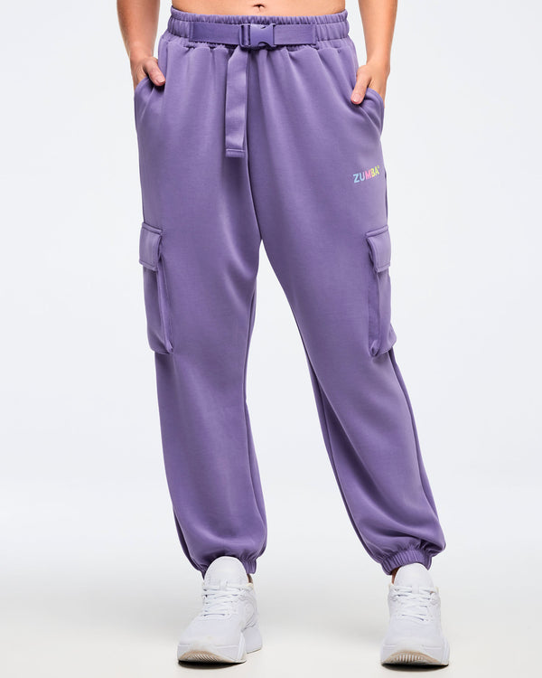 Front view of a female model wearing purple slouch cargo jogger pants with an elastic waistband, adjustable belt, side flap cargo pockets, and a small “Zumba” logo, paired with white sneakers on a white background.