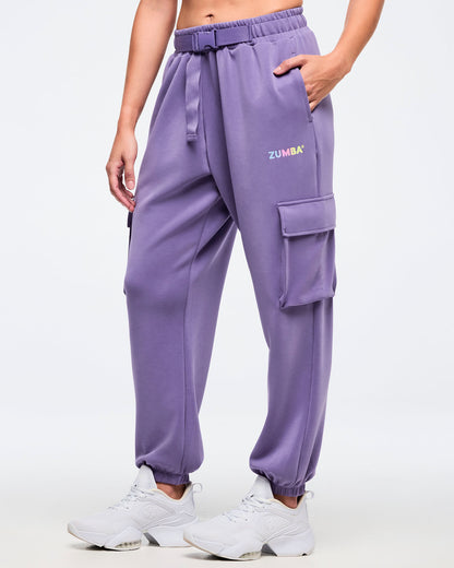 Close-up of purple slouch cargo jogger pants with an elastic waist and belt, side cargo pocket, and a small multicolor “ZUMBA” logo, paired with white sneakers on a white background.