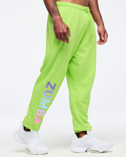 Neon green Zumba joggers with elastic drawstring waistband and tapered legs, featuring large vertical “ZUMBA” gradient lettering on the lower leg, worn by a male model with white sneakers.