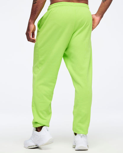 Back angled view of neon green Zumba joggers with elastic waistband and tapered legs, worn by a male model with white sneakers.