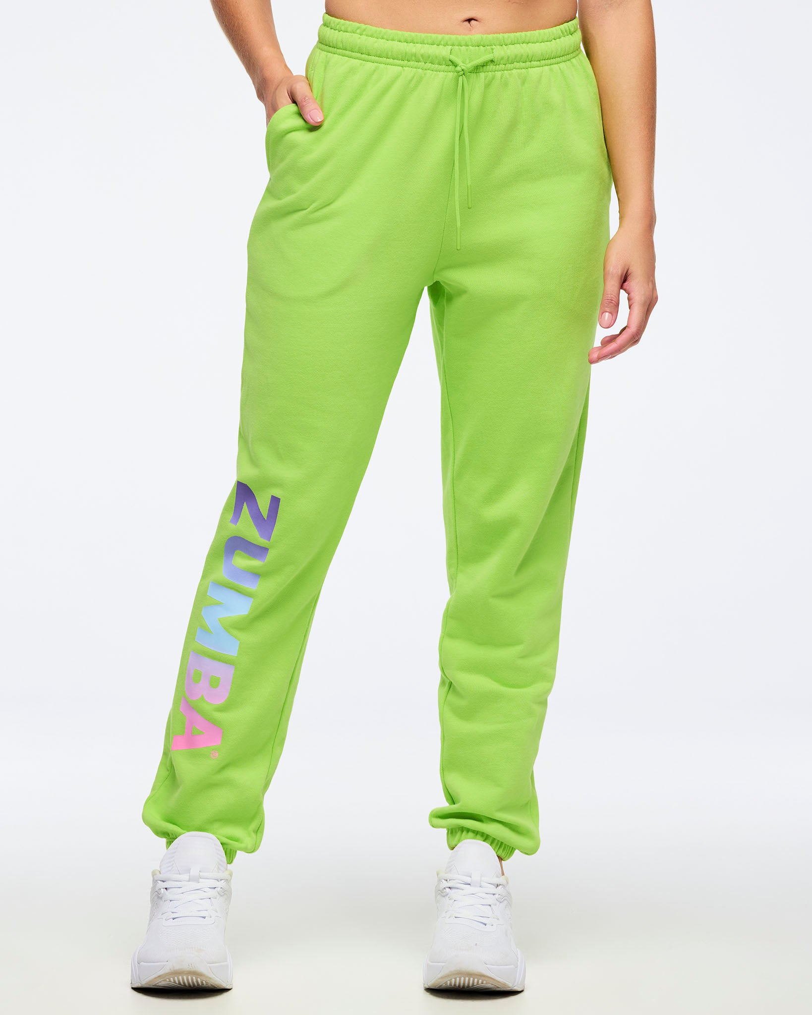 Front view of neon green Zumba joggers with elastic drawstring waistband and side pockets, featuring large vertical “ZUMBA” gradient lettering on the leg, worn by a female model with white sneakers.