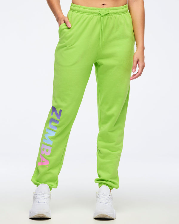 Front view of neon green Zumba joggers with elastic drawstring waistband and side pockets, featuring large vertical “ZUMBA” gradient lettering on the leg, worn by a female model with white sneakers.