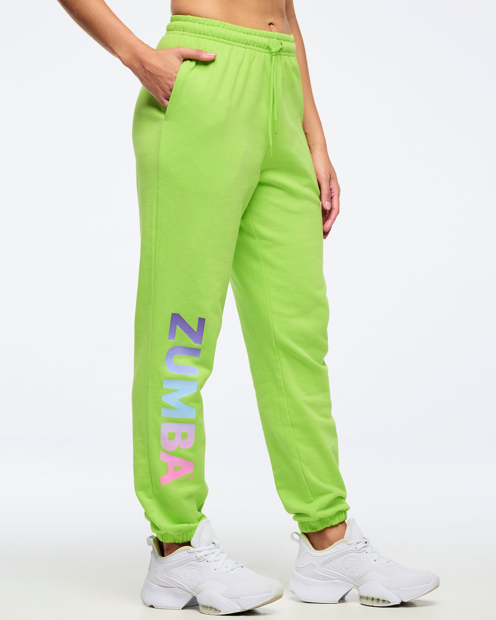 Neon green Zumba joggers with elastic drawstring waistband, side pockets, and ankle cuffs, featuring large vertical “ZUMBA” gradient lettering on the leg, worn by a female model with white sneakers.