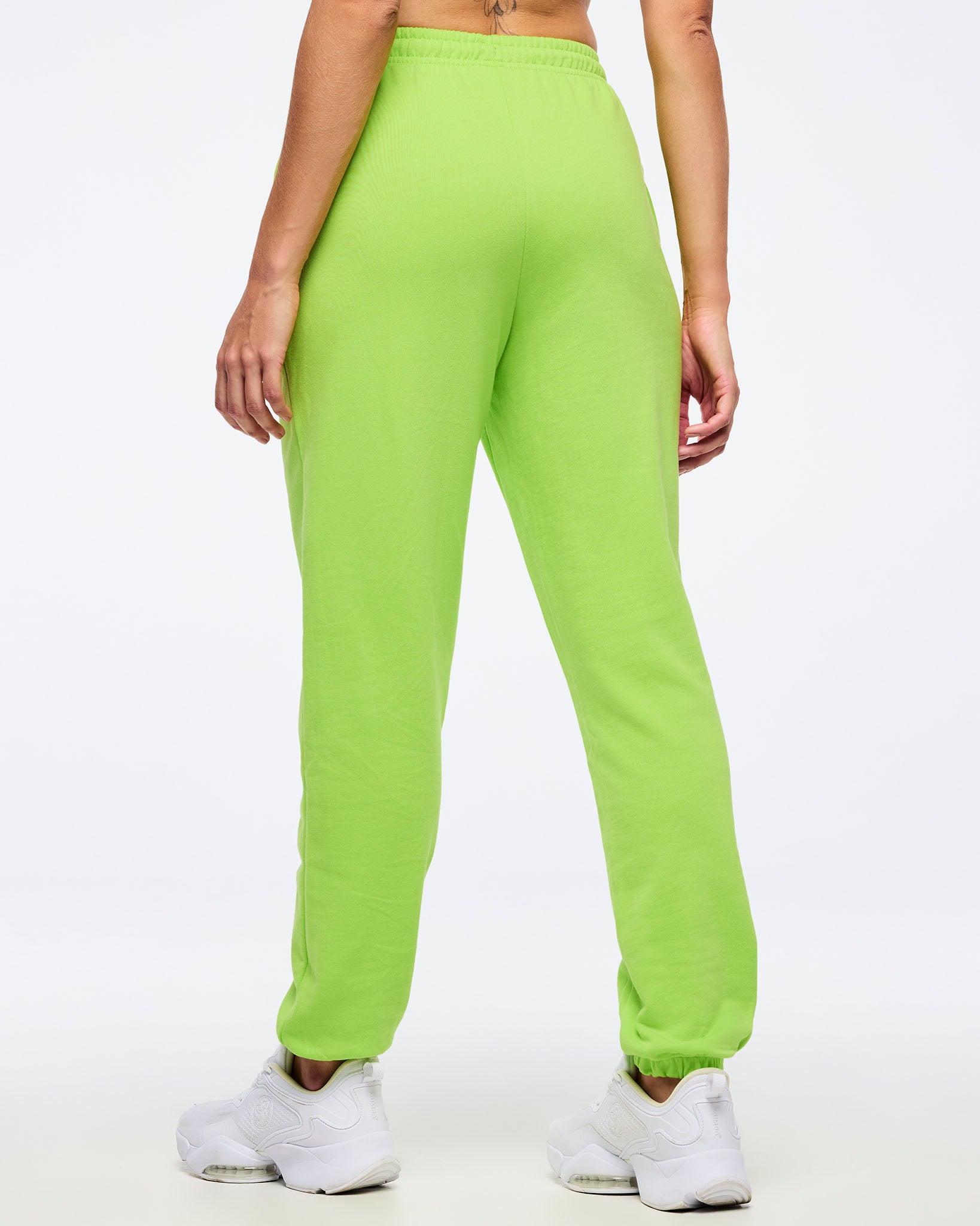 Back view of neon green Zumba joggers with elastic waistband and tapered ankles, worn by a female model with white sneakers.