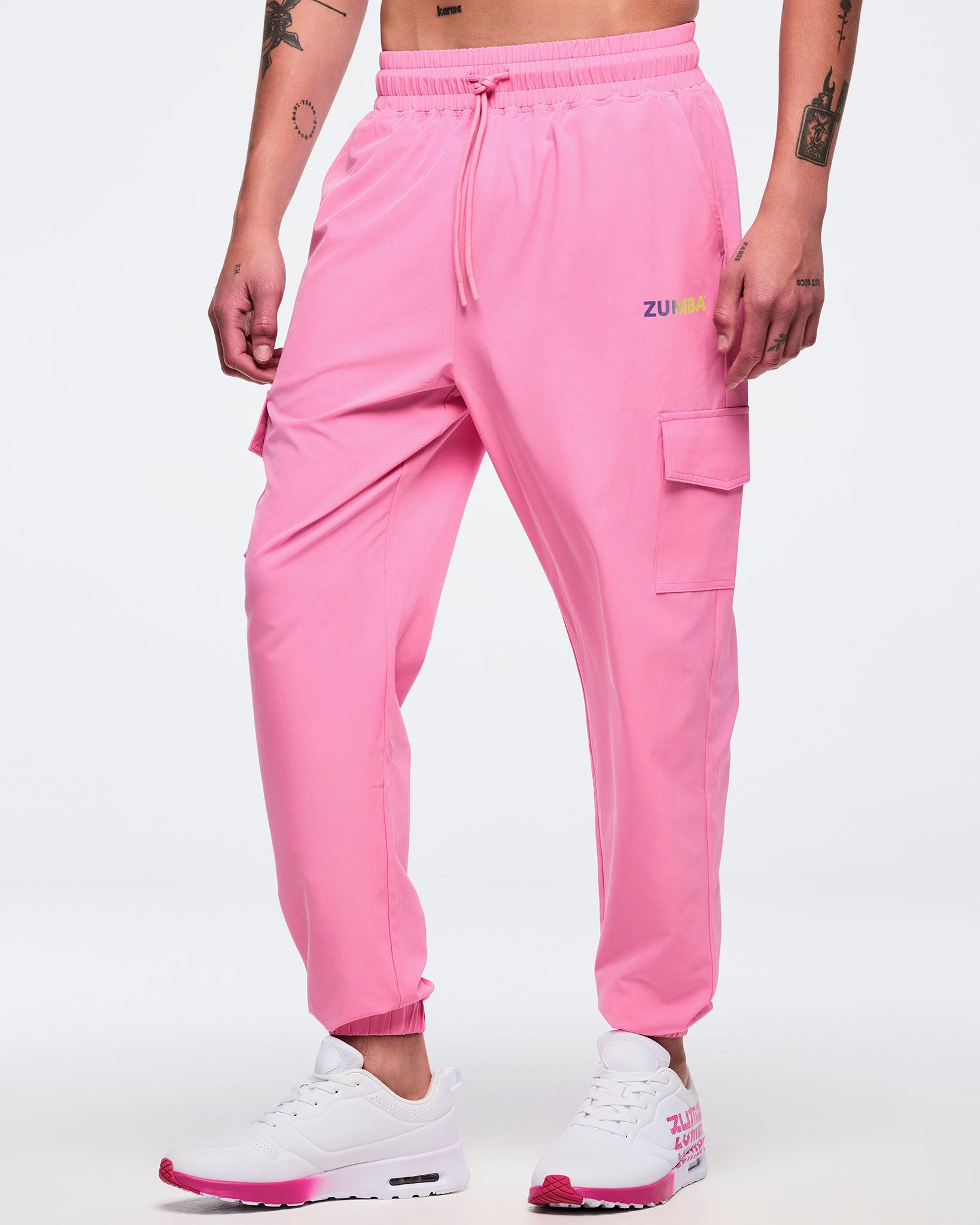 Front view of pink Zumba cargo joggers with elastic waistband and drawcord, side pockets, and a thigh flap cargo pocket, worn by a male model with white sneakers and pink soles.