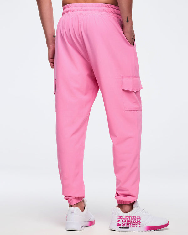 Back view of pink Zumba cargo joggers with elastic waistband and ankle cuffs, showing a flap cargo pocket on the thigh, worn by a male model with white-and-pink sneakers.