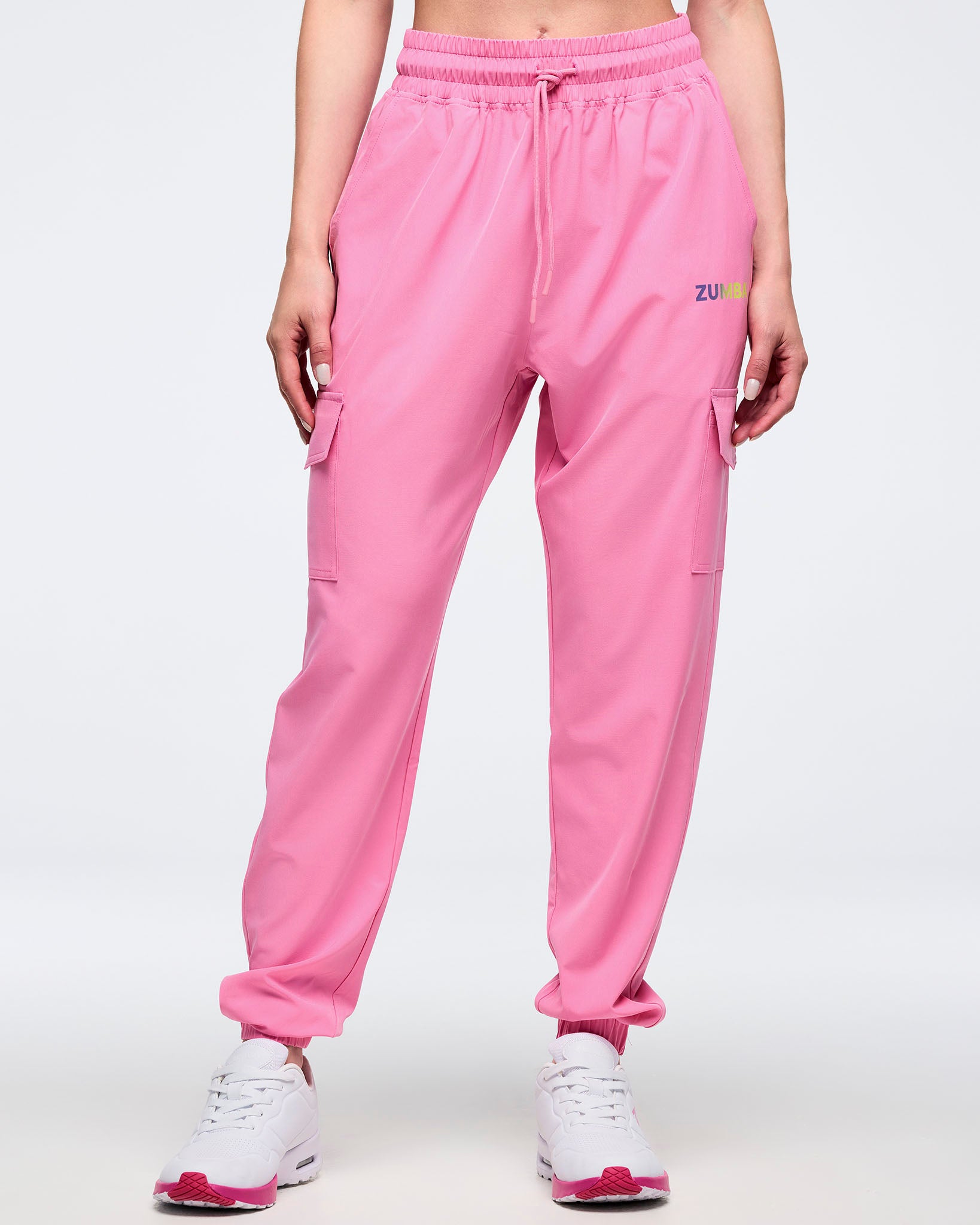 Full front view of pink Zumba cargo joggers with elastic waistband and drawcord, flap cargo pockets on both thighs, and tapered ankles, worn by a female model with white sneakers and pink soles.