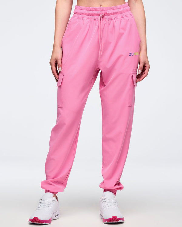 Full front view of pink Zumba cargo joggers with elastic waistband and drawcord, flap cargo pockets on both thighs, and tapered ankles, worn by a female model with white sneakers and pink soles.