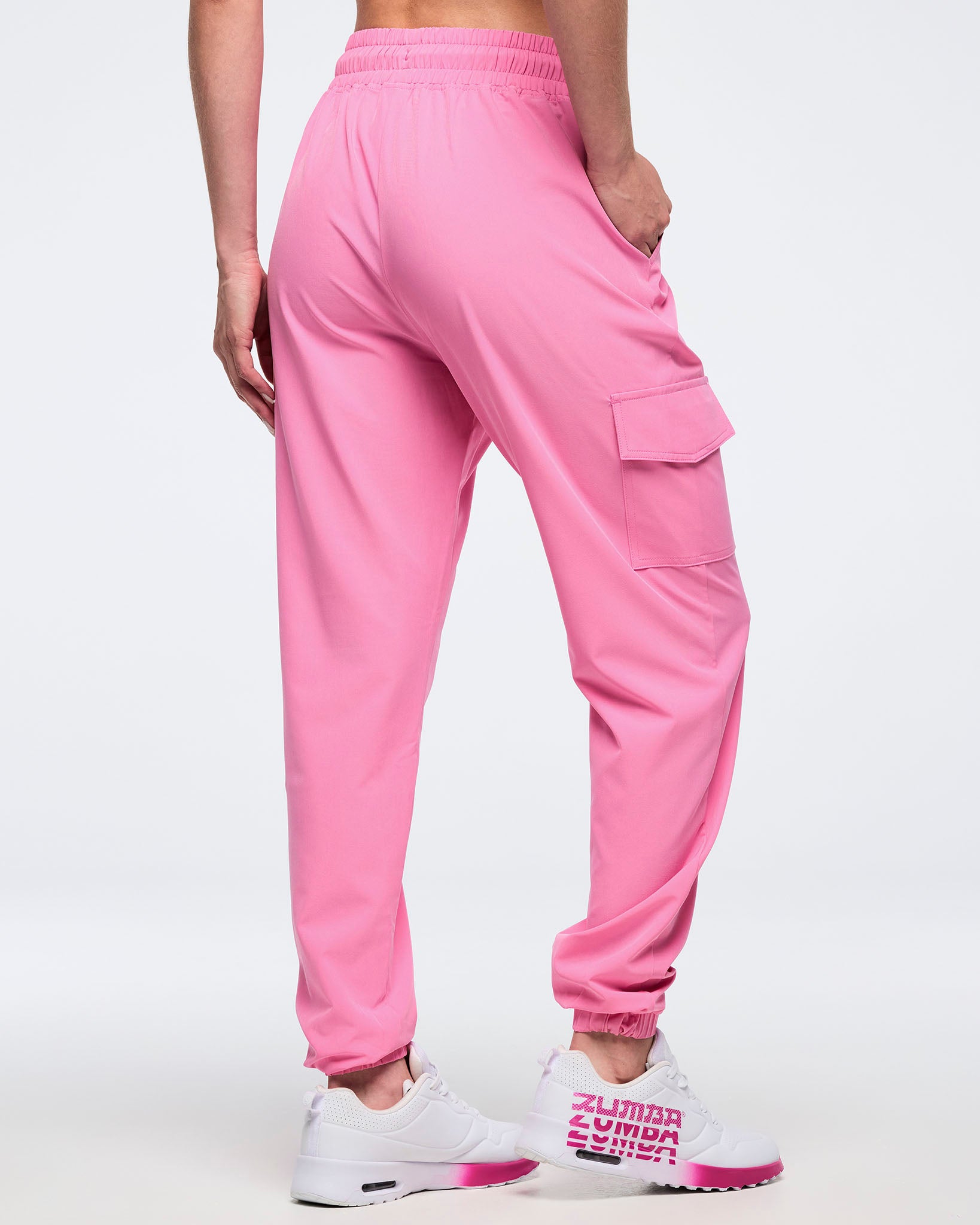 Back angled view of pink Zumba cargo joggers with elastic waistband, ankle cuffs, and a thigh flap cargo pocket, worn by a female model with white-and-pink sneakers.