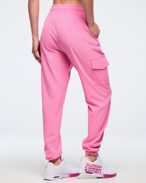 Back angled view of pink Zumba cargo joggers with elastic waistband, ankle cuffs, and a thigh flap cargo pocket, worn by a female model with white-and-pink sneakers.