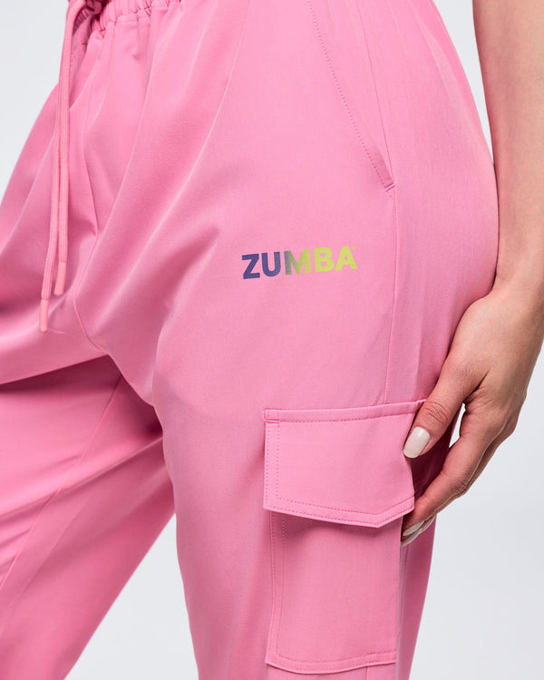 Close-up of pink pastel Zumba cargo joggers showing the small gradient “ZUMBA” logo, elastic waistband with drawcord, and a flap cargo pocket on the thigh, worn by a female model.