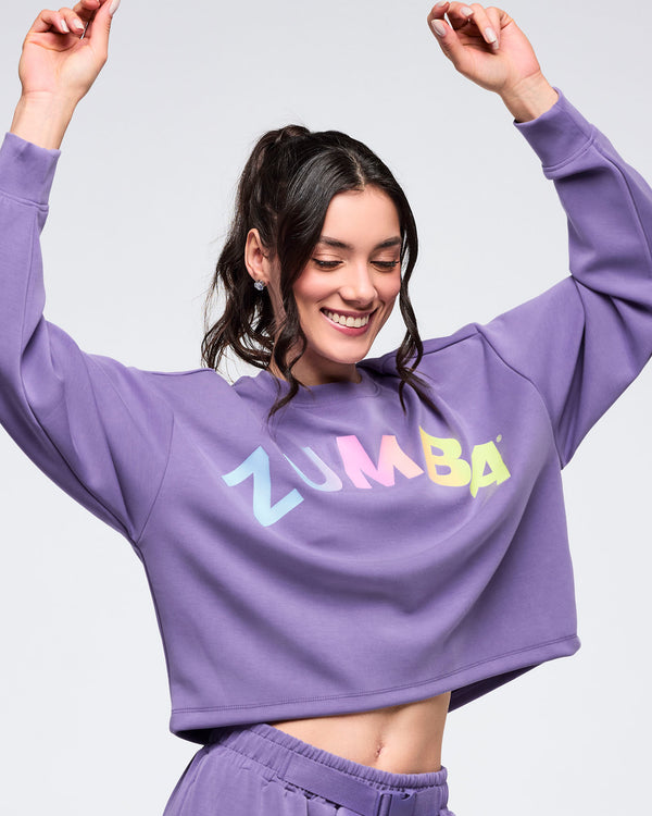 Purple “ZUMBA” cropped sweatshirt with multicolor gradient lettering, relaxed fit, and long sleeves, worn by a female model with matching purple joggers and arms raised against a light studio background.