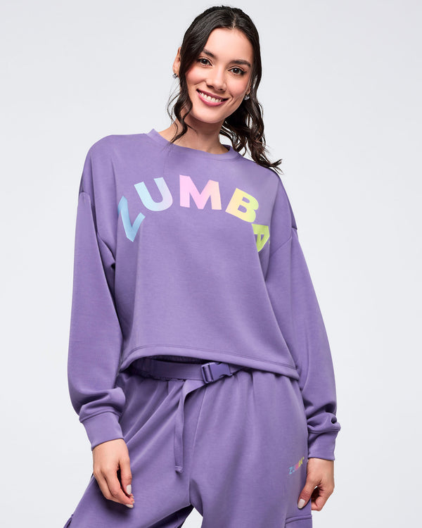 Close-up of a purple “ZUMBA” cropped sweatshirt with multicolor gradient lettering, crewneck, and long sleeves, worn by a female model with matching purple joggers against a light studio background.