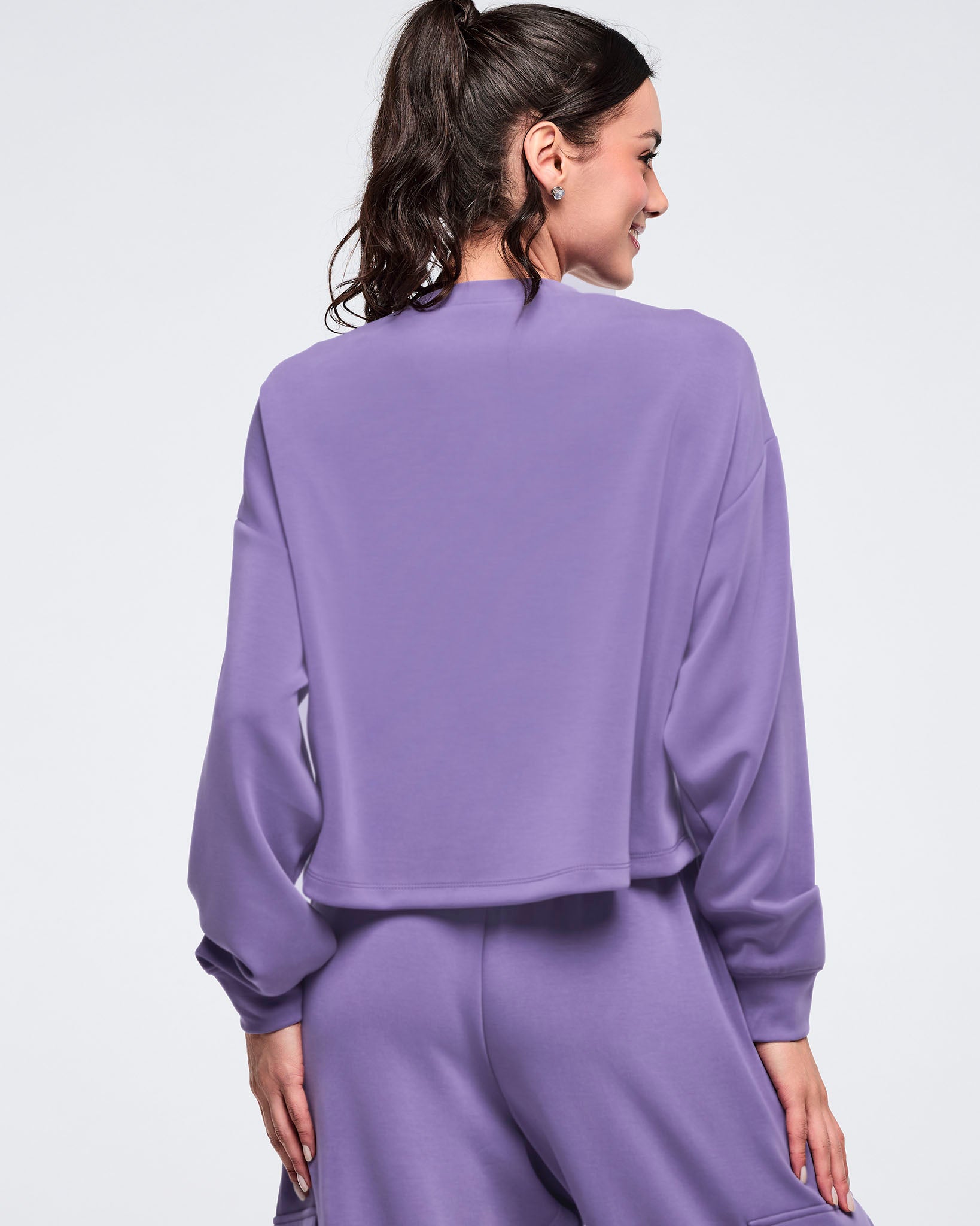 Back view of a purple cropped sweatshirt with a relaxed fit, dropped shoulders, and ribbed cuffs, worn by a female model with matching purple joggers against a light studio background.