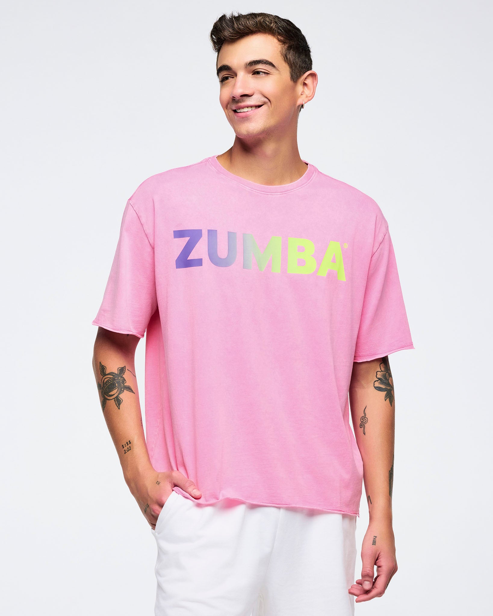 Pink pastel “ZUMBA” logo T-shirt with multicolor gradient lettering, relaxed fit and crewneck, worn by a male model with white shorts against a light studio background.