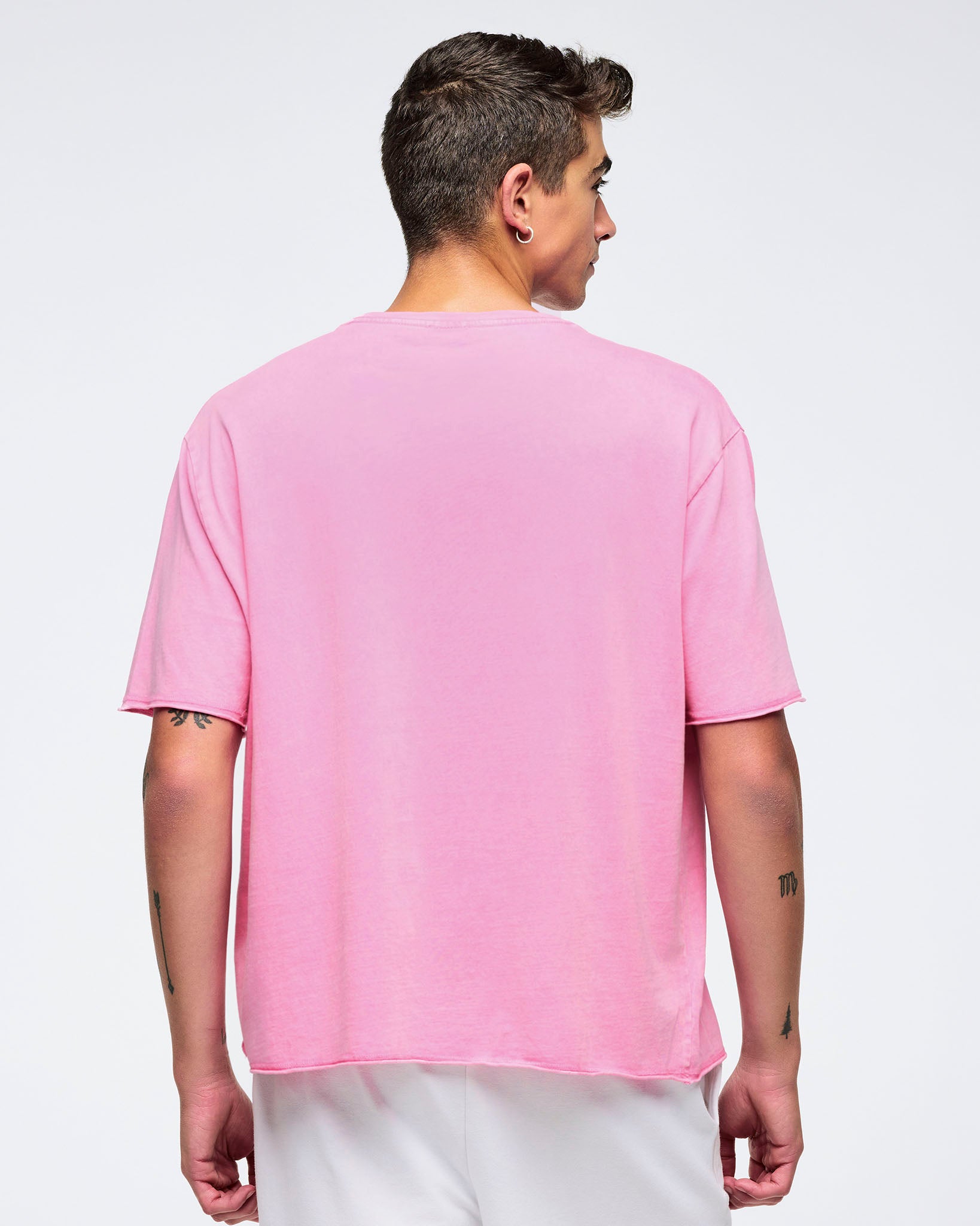 Back view of a pink pastel relaxed-fit crewneck T-shirt with dropped shoulders and elbow-length sleeves, worn by a male model with white shorts against a light studio background.