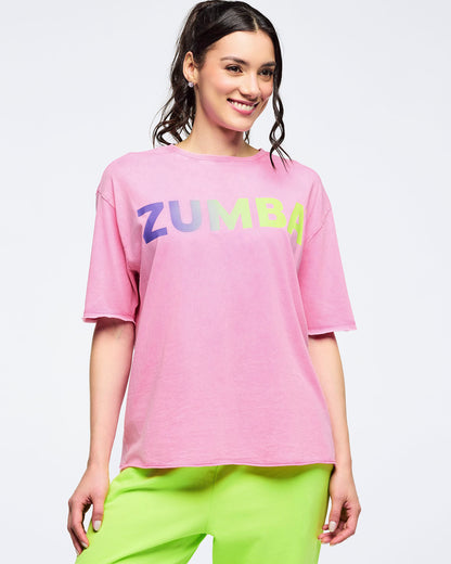 Pink pastel “ZUMBA” logo T-shirt with gradient lettering, relaxed fit and crewneck, worn by a female model with neon green pants against a light studio background.