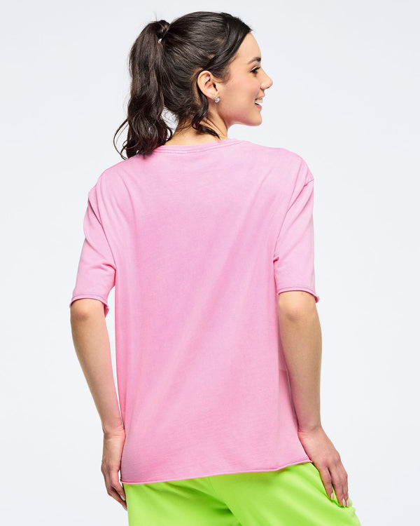 Back view of a pink pastel relaxed-fit crewneck T-shirt with elbow-length sleeves, worn by a female model with neon green pants against a light studio background.