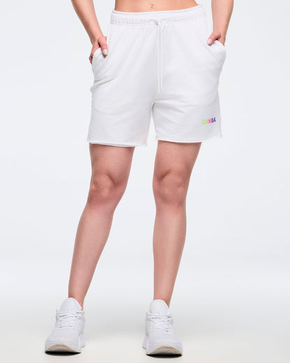 Woman in white shorts, with hands in her pockets. Zumba text in pastel colors on the left leg and the model on a white background.