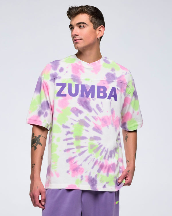 Man standing against a white background, looking to the side, wearing an oversized pastel tie-dye pattern T-shirt with a large purple “ZUMBA” logo and purple pants.